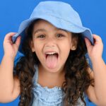 Menina sorridente mostrando a língua e usando chapéu azul em fundo azul