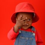 Menino sorridente com chapéu vermelho cobrindo parte do rosto em fundo vermelho